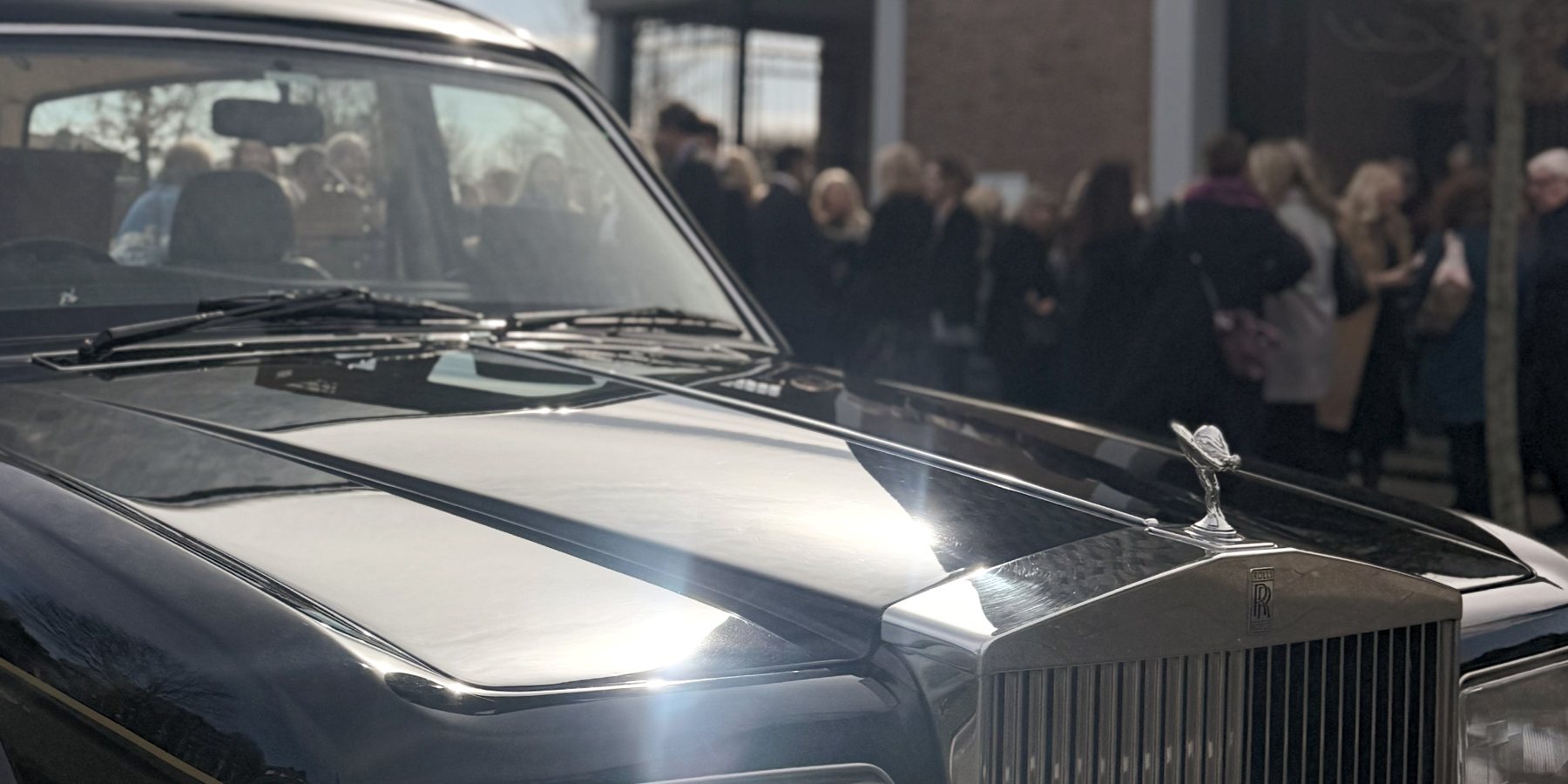 The bonnet of a Rolls Royce hearse with mourners gathered behind