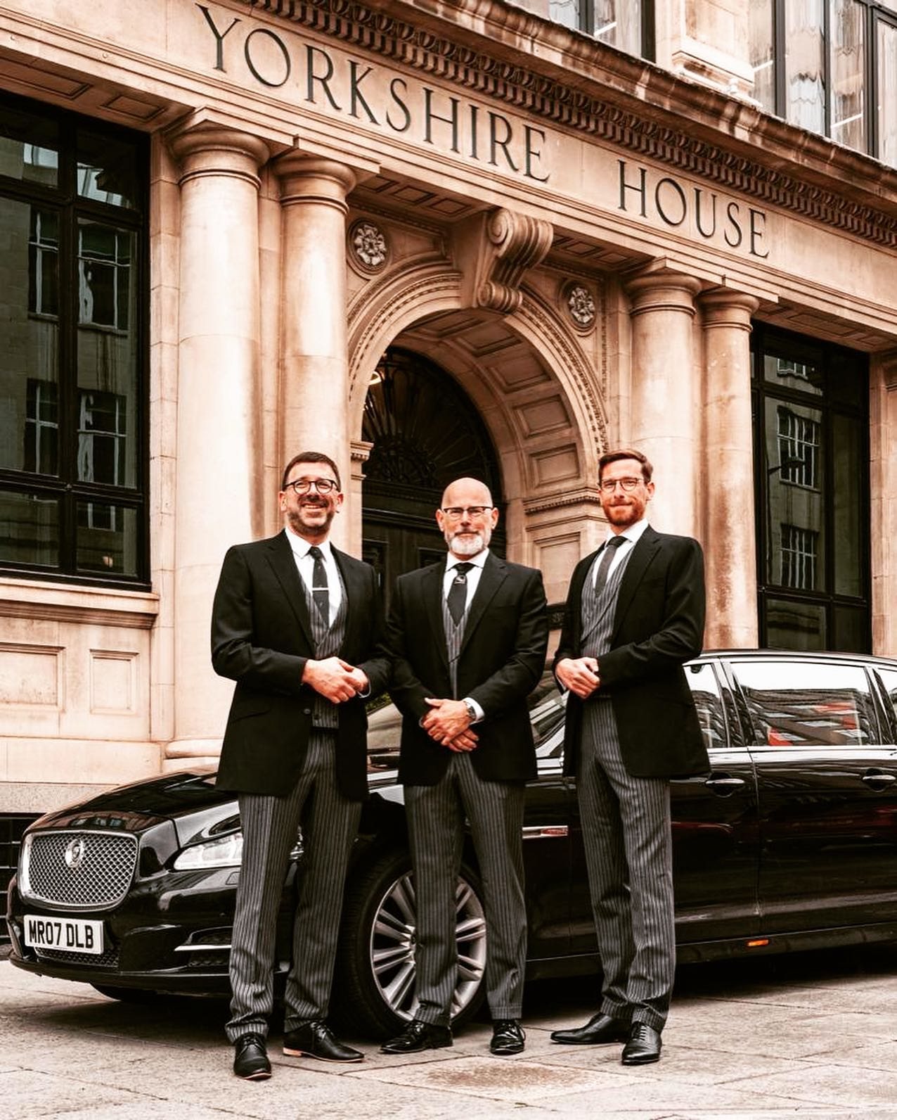 Three men in dark suits standing beside a Jaguar limousine
